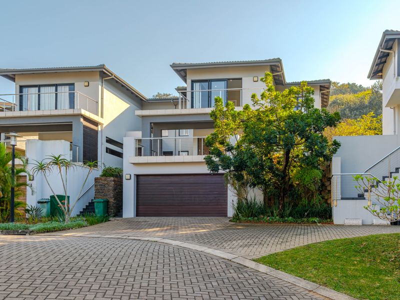 Modern townhouse exterior featuring a double garage, paved driveway, and upper-level balconies with stainless steel balustrades, set in a well-maintained residential complex.
