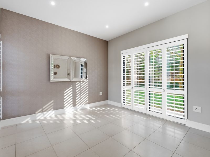 Bright and spacious living area featuring neutral tiled flooring, elegant textured wallpaper, and white plantation shutters opening onto the garden.