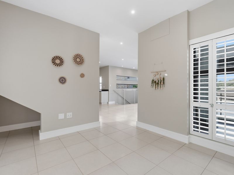 A bright, modern hallway featuring neutral tiled flooring, recessed lighting, and white plantation shutters, leading towards an open-plan living area.