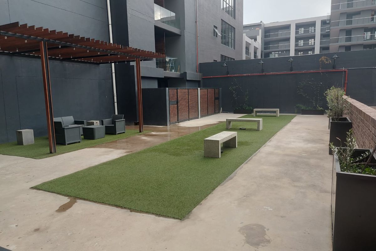 A modern communal outdoor courtyard featuring a timber pergola, artificial lawn areas, and built-in concrete seating within an apartment complex.