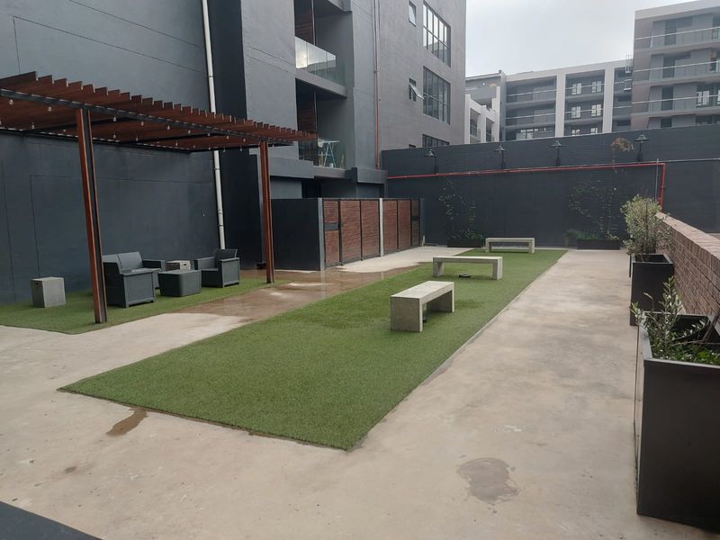A modern communal outdoor courtyard featuring a timber pergola, artificial lawn areas, and built-in concrete seating within an apartment complex.