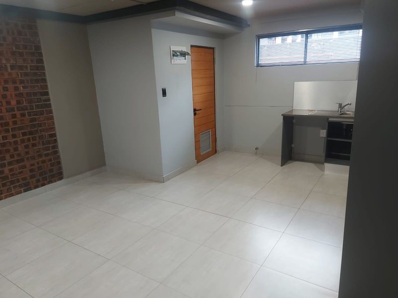 An open-plan living area featuring tiled floors, an exposed brick accent wall, and a compact kitchenette with sink and cabinetry.