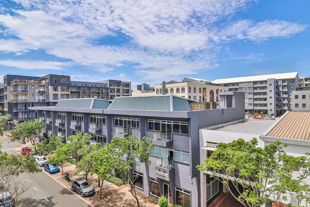 An elevated aerial view of a modern multi-story apartment complex featuring grey facades, metal roofing, and private balconies overlooking a tree-lined street.