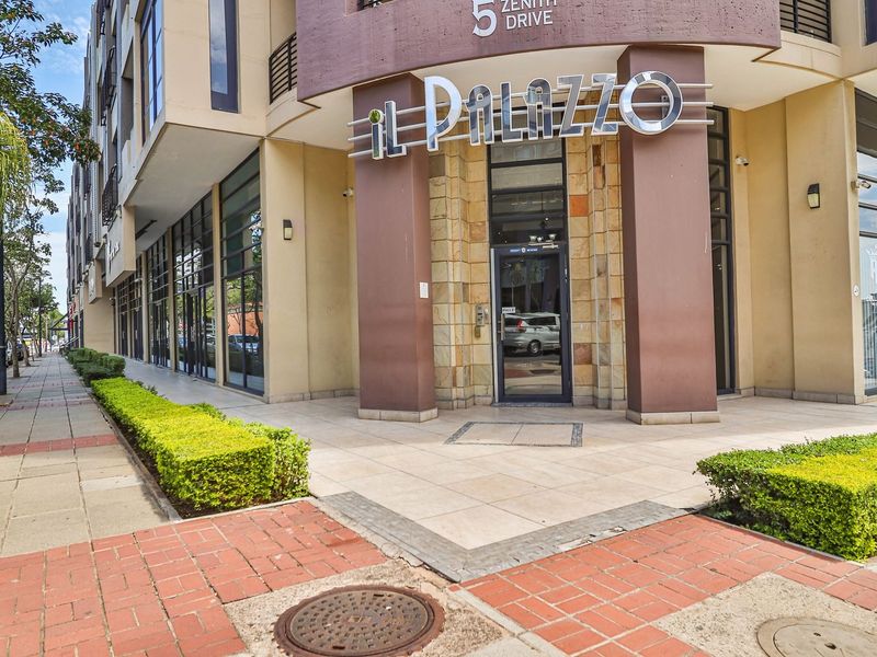 The front entrance of Il Palazzo apartment building, featuring stone cladding, a glass door, and well-maintained paved walkways with landscaped hedging.