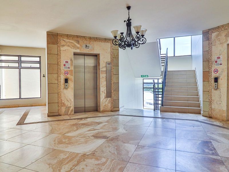 A spacious apartment building lobby featuring marble flooring, dual elevators, and a central staircase with stone-clad accents.