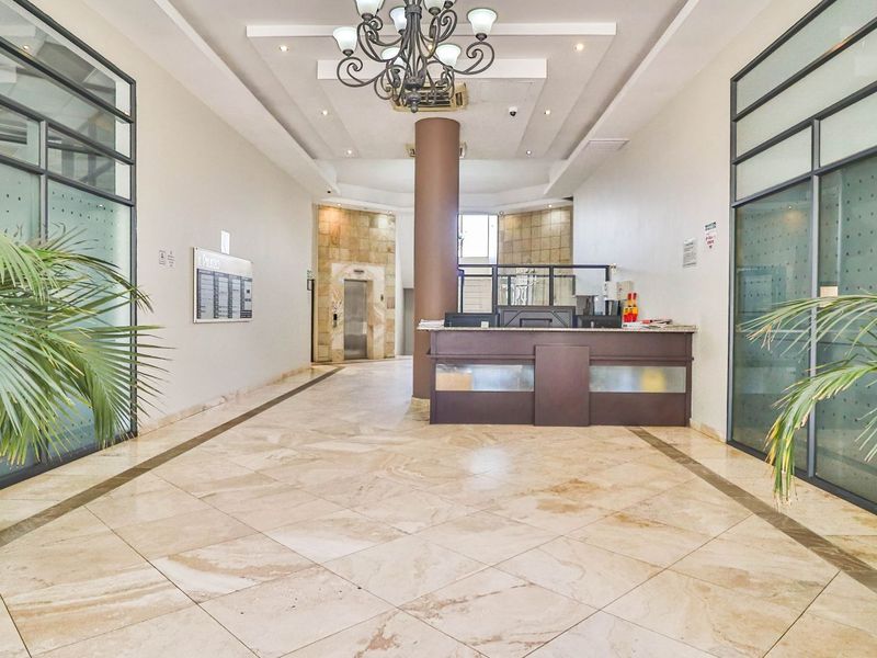 A spacious and well-maintained apartment building lobby featuring a reception desk, elevator access, tiled flooring, and modern glass partition walls.