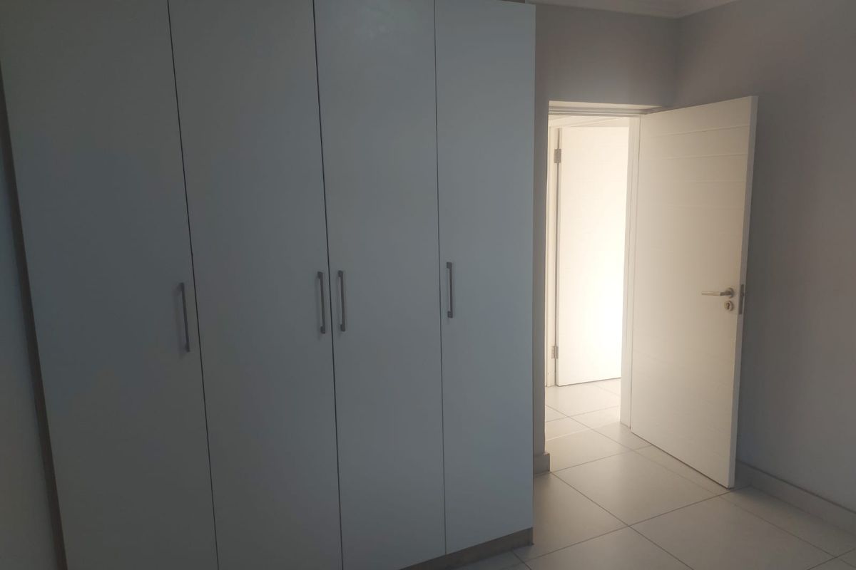 A clean, empty bedroom featuring modern built-in cupboards, neutral tiled flooring, and a bright, open doorway leading to an adjacent space.
