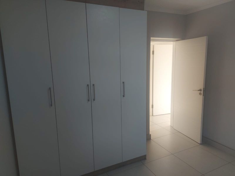 A clean, empty bedroom featuring modern built-in cupboards, neutral tiled flooring, and a bright, open doorway leading to an adjacent space.