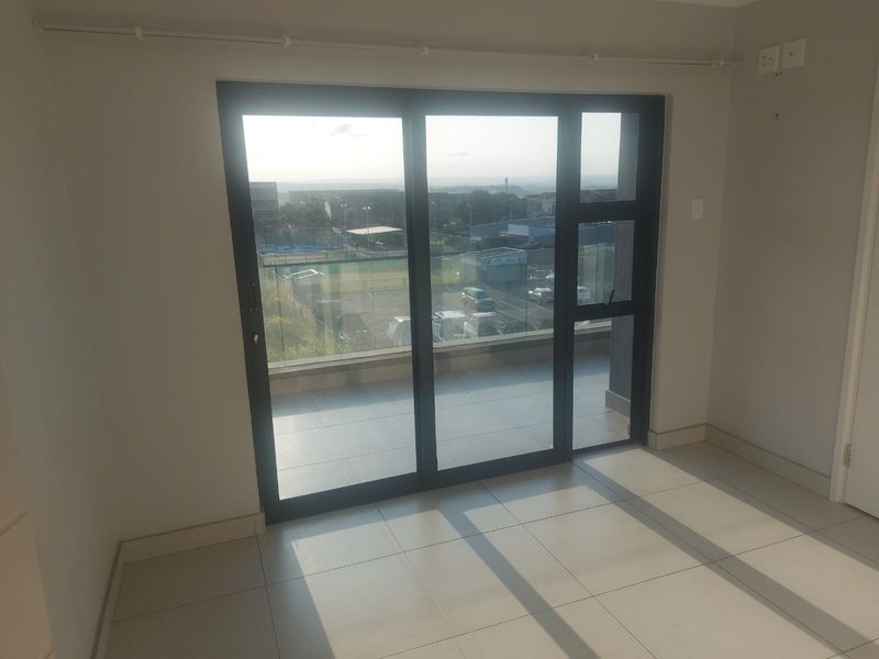 An empty, well-lit living area featuring neutral tiled flooring and large sliding glass doors that provide access to a balcony with exterior views.