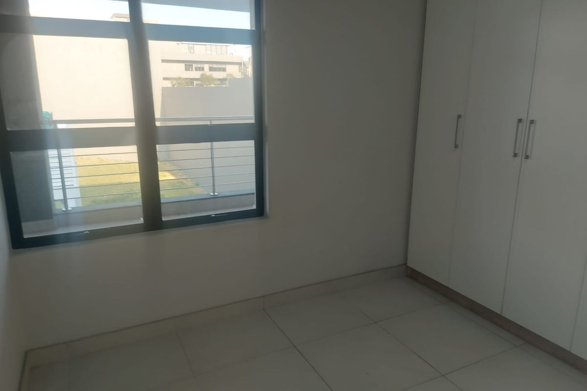 A bright, empty bedroom featuring neutral tiled flooring, built-in white cupboards, and large aluminium-framed windows providing natural light.