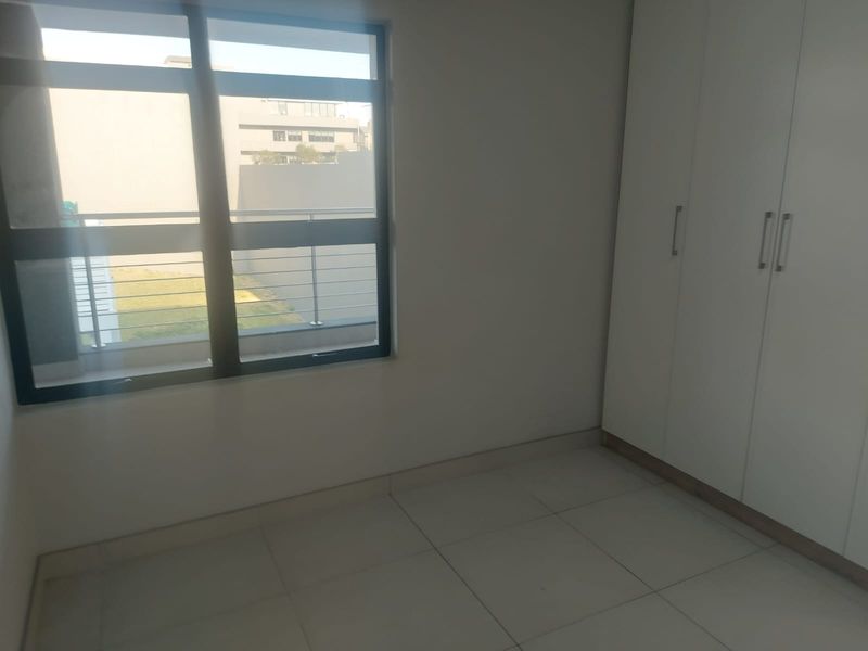A bright, empty bedroom featuring neutral tiled flooring, built-in white cupboards, and large aluminium-framed windows providing natural light.