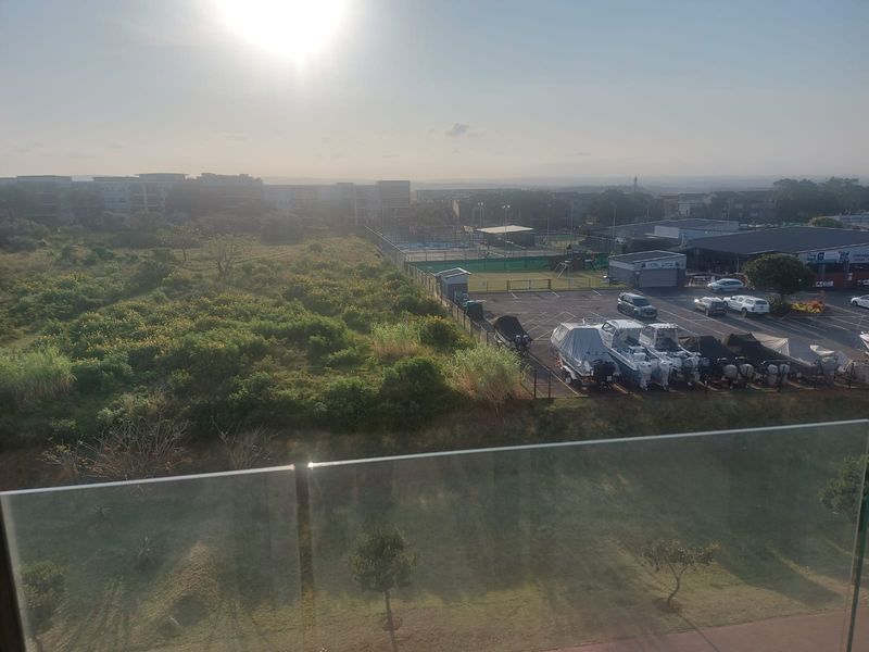 Elevated view overlooking a landscaped area, parking lot, and surrounding neighborhood, captured from a balcony with a modern glass balustrade.