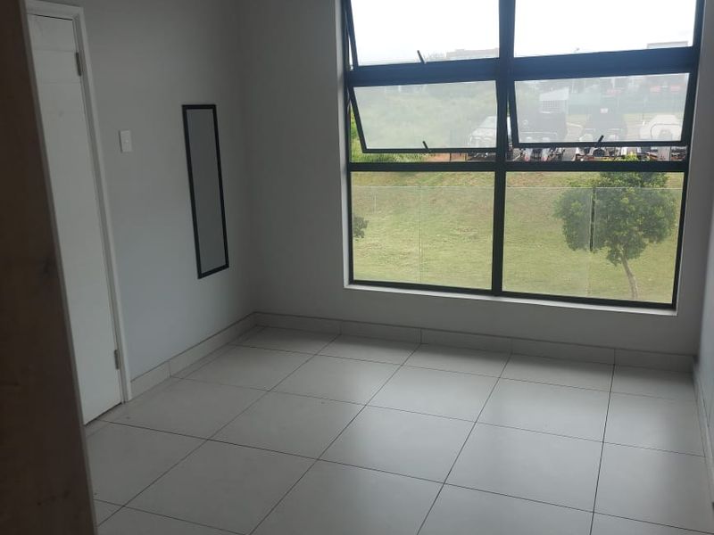 A bright, empty bedroom featuring neutral tiled flooring and a large window with a view of the surrounding green space.