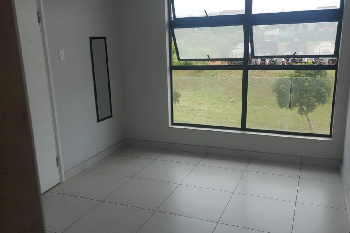 A bright, empty bedroom featuring neutral tiled flooring and a large window with a view of the surrounding green space.