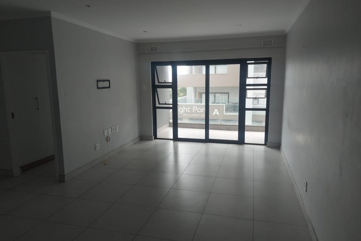 An empty, open-plan living area featuring neutral tiled flooring, recessed lighting, and large sliding glass doors leading to a balcony.