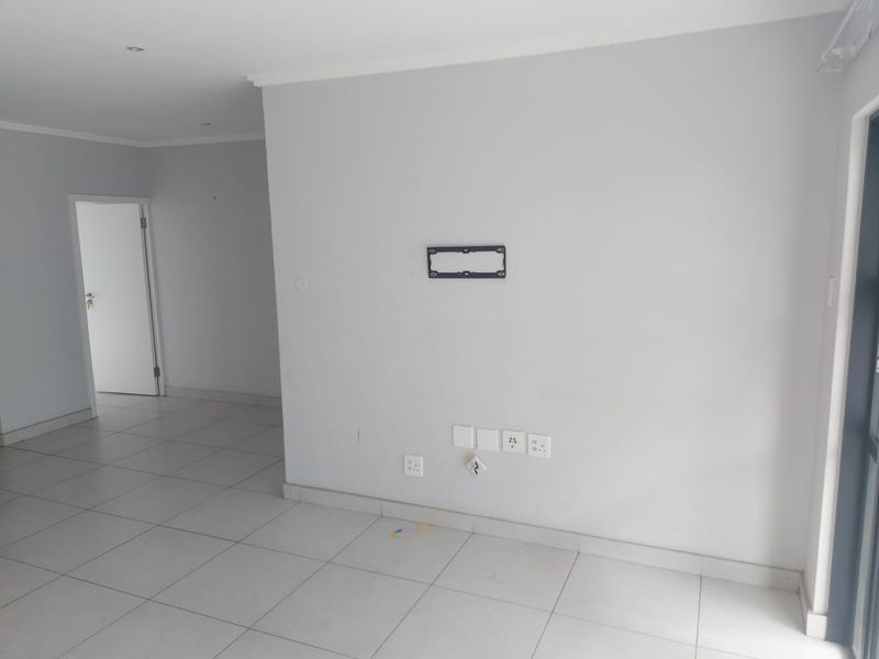 A bright, empty living area featuring neutral tiled flooring, recessed lighting, and a wall-mounted TV bracket with integrated electrical and data points.