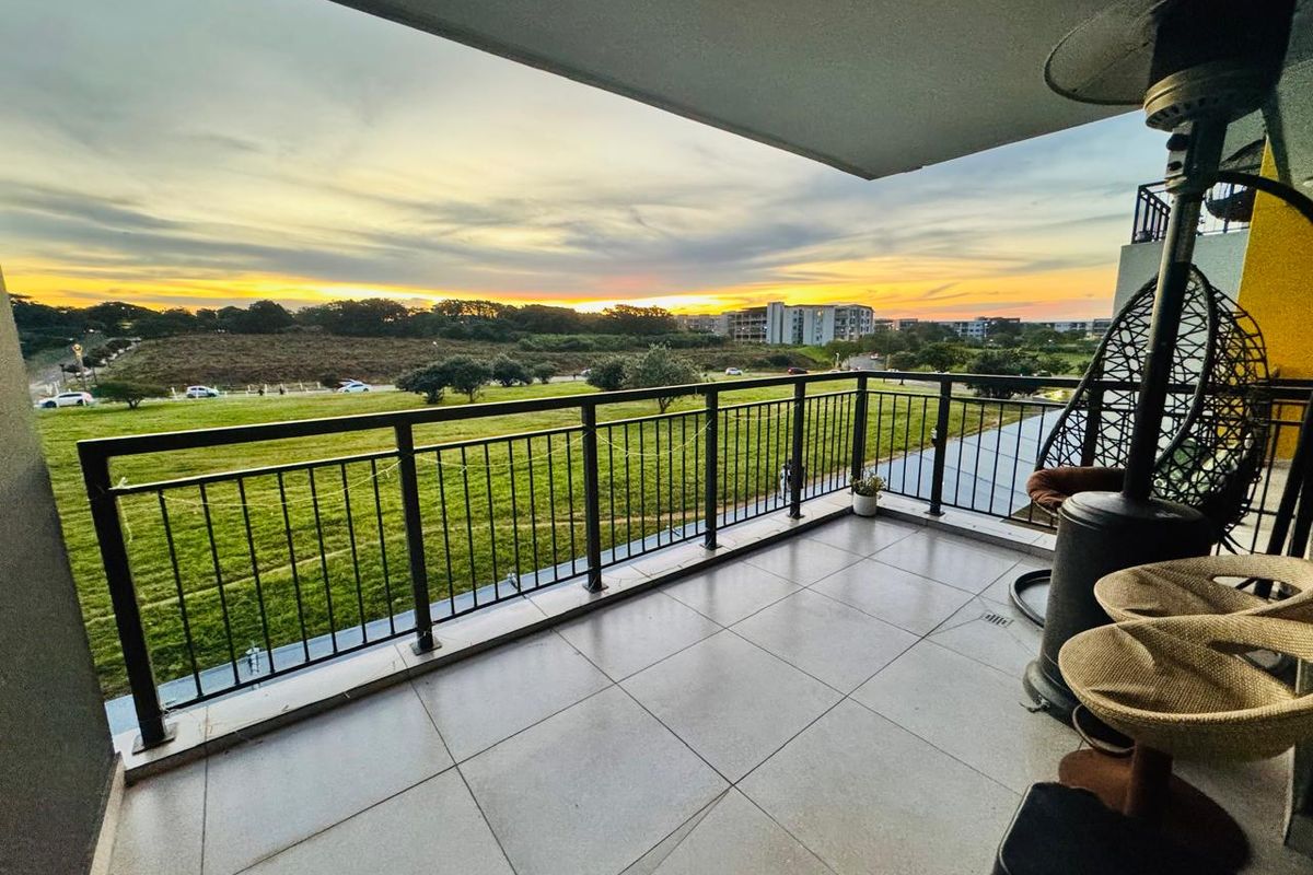 A spacious covered balcony featuring tiled flooring and a metal balustrade, offering scenic sunset views over the surrounding green landscape.