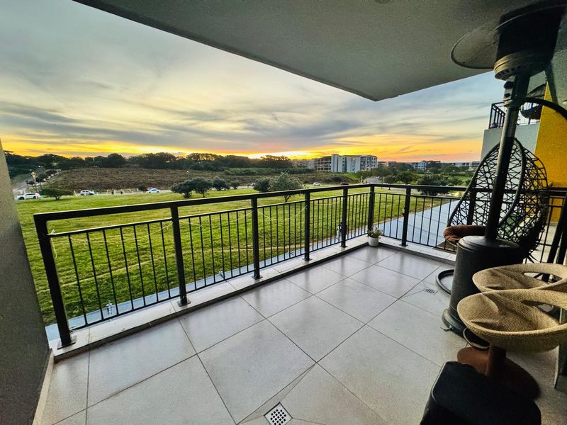 A spacious covered balcony featuring tiled flooring and a metal balustrade, offering scenic sunset views over the surrounding green landscape.
