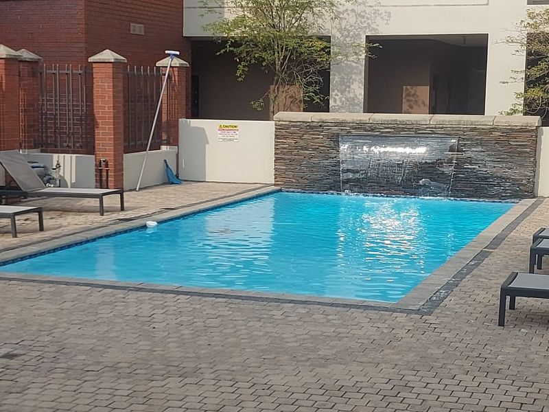 Communal swimming pool area featuring a stone-clad water feature and paved decking, set within a well-maintained apartment complex courtyard.