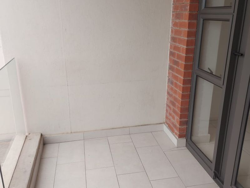A clean, modern balcony featuring light-toned floor tiles, a glass balustrade, and an exposed brick accent pillar with an aluminium-framed glass door.
