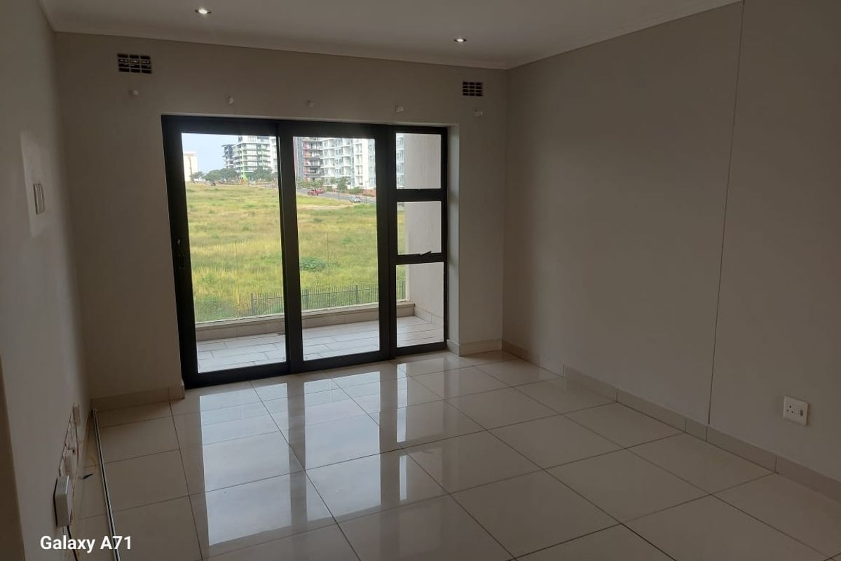 A bright, empty living area featuring large sliding glass doors that open onto a balcony, with neutral tiled flooring and recessed lighting.