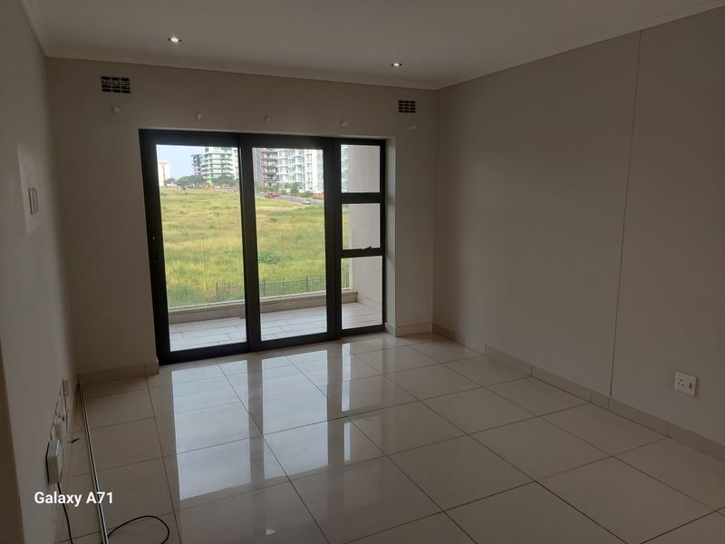 A bright, empty living area featuring large sliding glass doors that open onto a balcony, with neutral tiled flooring and recessed lighting.
