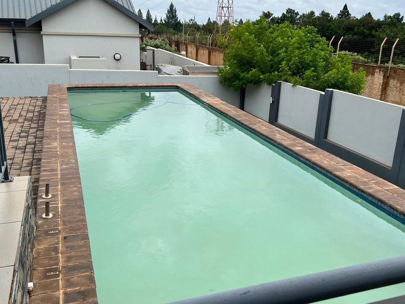 A rectangular swimming pool with brick coping, surrounded by a paved patio area and enclosed by a perimeter wall and metal safety railing.