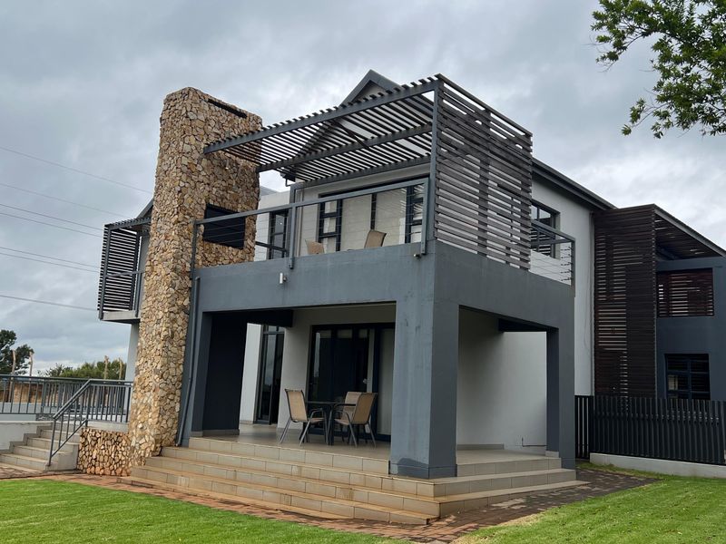 Rear elevation of a modern house featuring a stone-clad chimney, a covered patio, and an upper-level balcony with timber pergola detailing.