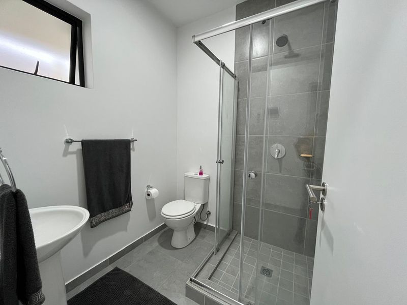 A modern, clean bathroom featuring a pedestal sink, a toilet, and a glass-enclosed shower with grey tiled finishes.