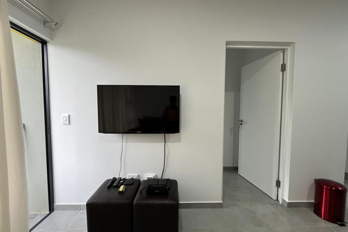 A simple living area featuring neutral tiled flooring, a wall-mounted television, and direct access to an outdoor space via a sliding glass door.