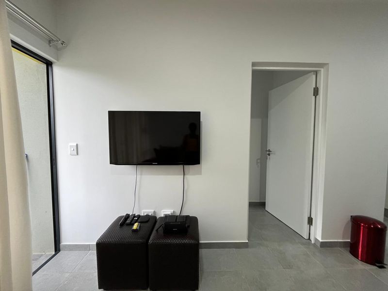 A simple living area featuring neutral tiled flooring, a wall-mounted television, and direct access to an outdoor space via a sliding glass door.