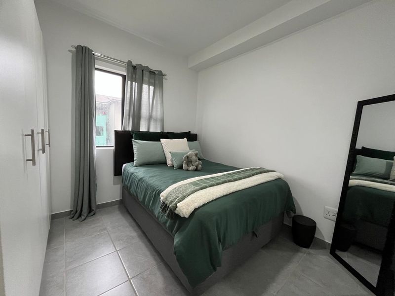A compact, well-maintained bedroom featuring neutral tiled flooring, a built-in wardrobe, and a window providing natural light.