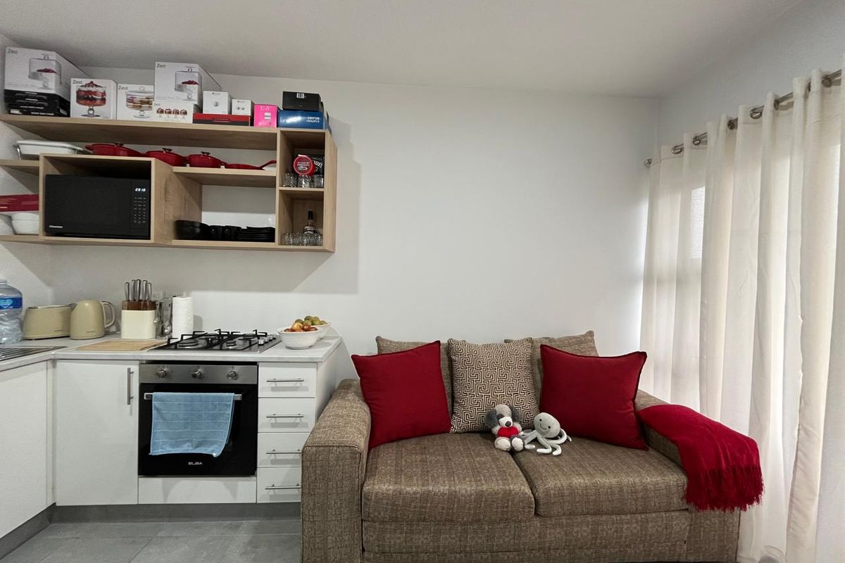 Compact open-plan living area featuring a modern kitchen corner with built-in oven, gas hob, and overhead shelving, adjacent to a bright window space.