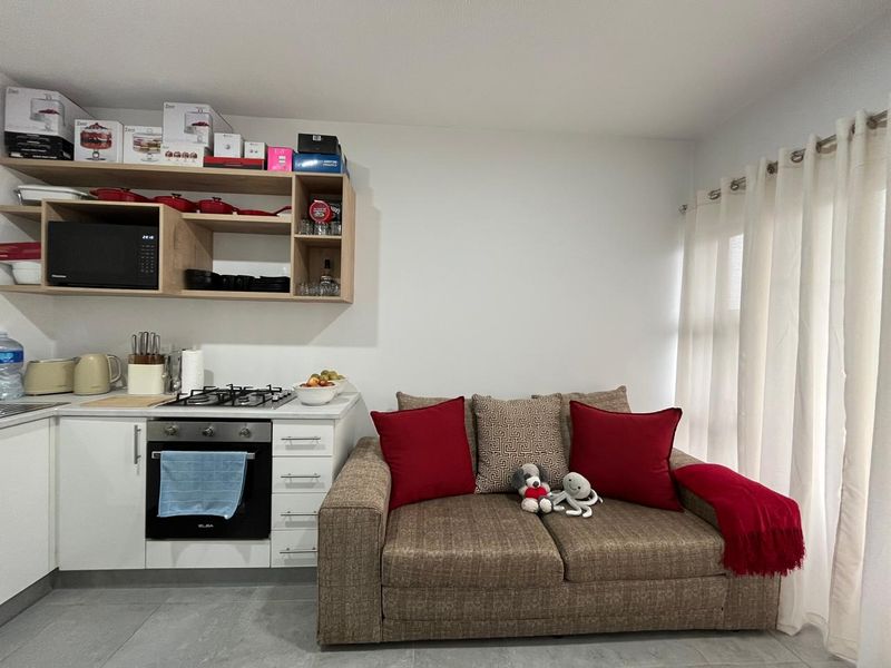 Compact open-plan living area featuring a modern kitchen corner with built-in oven, gas hob, and overhead shelving, adjacent to a bright window space.