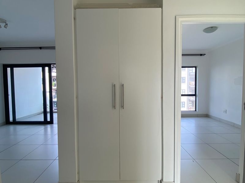 A bright, empty hallway featuring neutral tiled flooring, coved cornicing, and a central built-in storage cupboard, providing access to adjacent living areas.