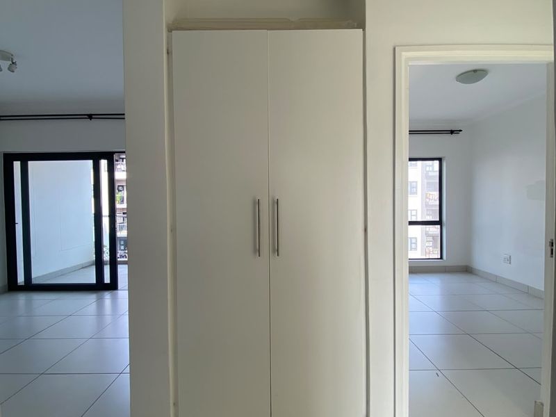 A bright, empty hallway featuring neutral tiled flooring, coving, and a central built-in cupboard providing convenient storage space.