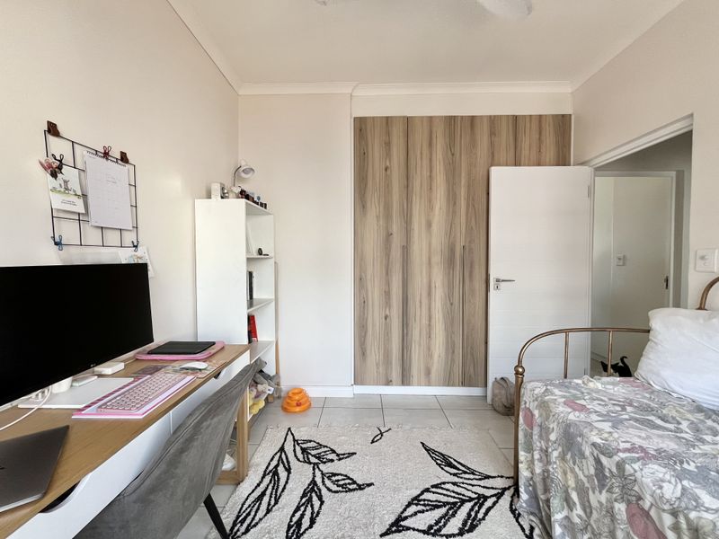 A well-maintained bedroom featuring modern wood-finish built-in cupboards, neutral tiled flooring, and a clean, functional layout suitable for a study or sleeping area.
