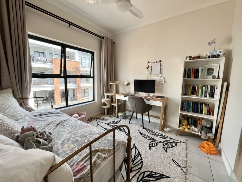 A well-lit bedroom featuring tiled flooring, a ceiling fan, and large aluminium-framed windows providing natural light and views of the surrounding apartment complex.