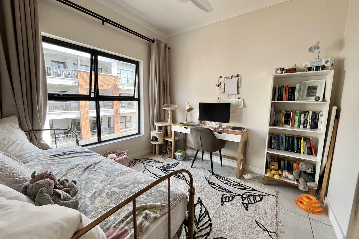 A well-lit bedroom featuring tiled flooring, a ceiling fan, and large aluminium-framed windows providing natural light and views of the surrounding apartment complex.