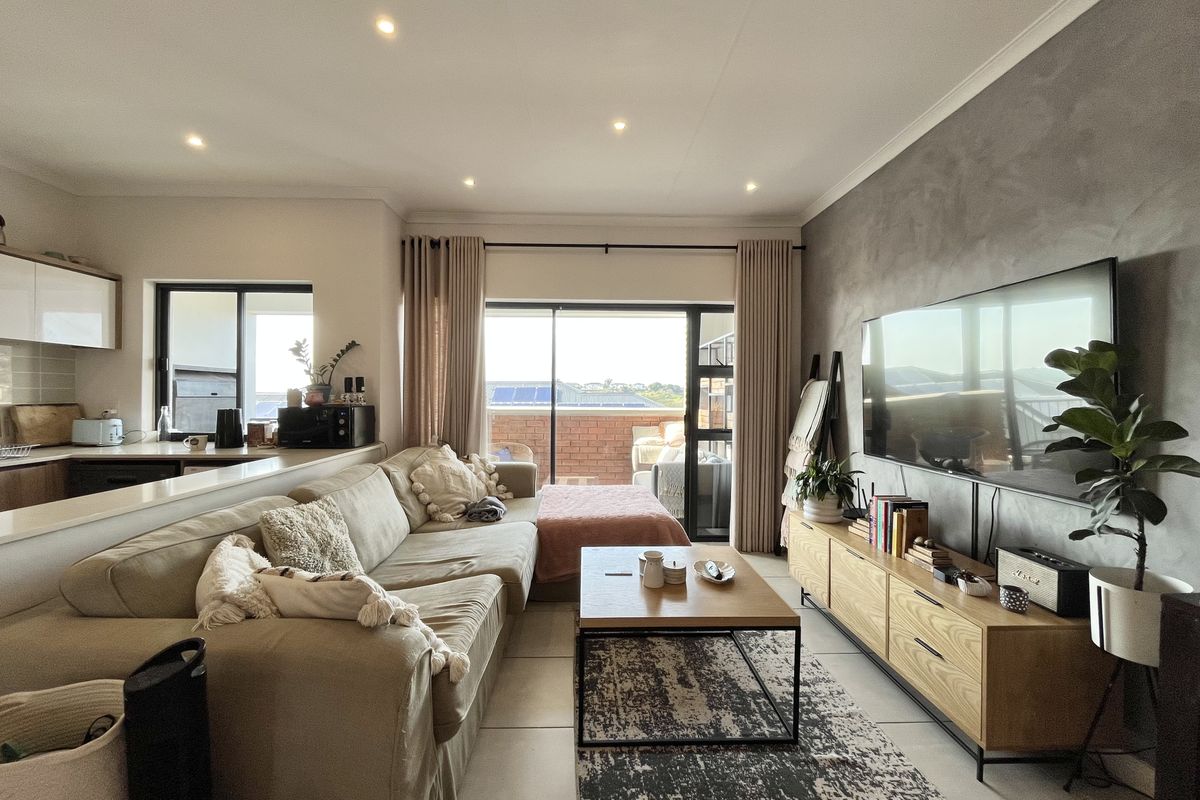 An open-plan living area featuring a modern kitchen, tiled floors, a textured feature wall, and direct access to a private balcony through sliding glass doors.