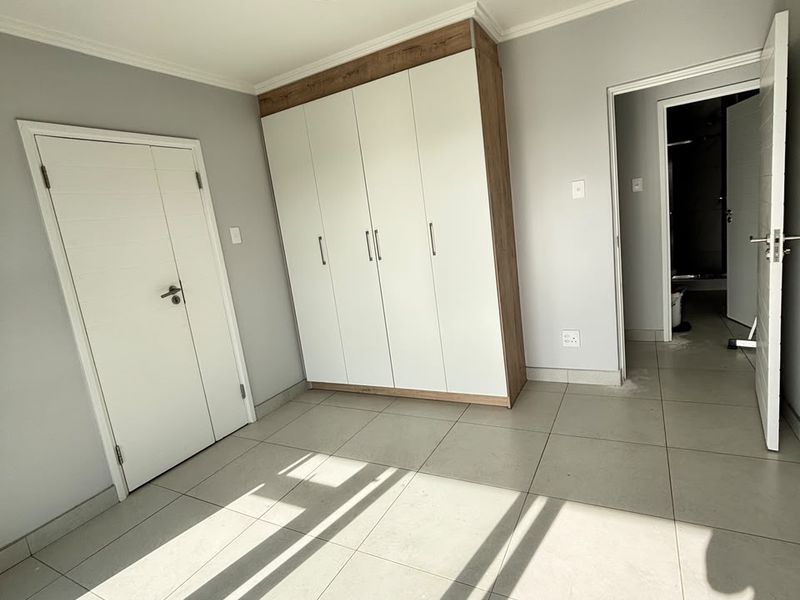 A bright, empty bedroom featuring neutral tiled flooring, a modern built-in wardrobe with wood-tone trim, and direct access to an en-suite bathroom.