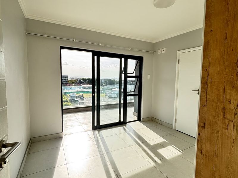 An empty, well-lit living area featuring large sliding glass doors that open onto a balcony, with neutral tiled flooring and modern finishes.