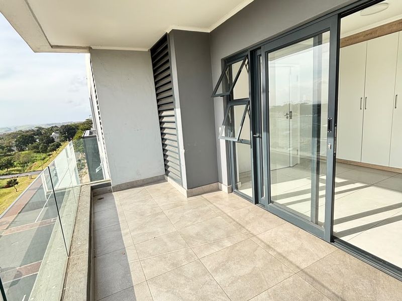 A modern, covered balcony featuring tiled flooring, a sleek glass balustrade, and sliding door access to the interior living space.