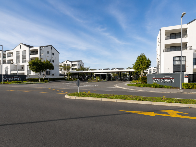 Street view of The Sandown apartment complex entrance, featuring modern white multi-story buildings, a secure gatehouse, and well-maintained paved access roads.