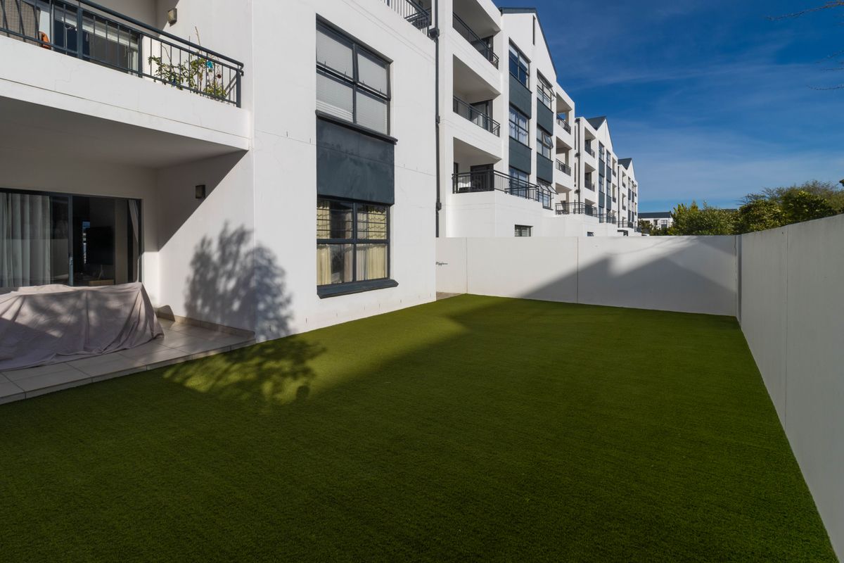 A private rear garden area featuring artificial turf, a paved patio, and direct access to the apartment via sliding glass doors.