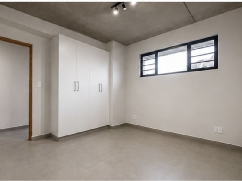 A clean, modern bedroom featuring built-in cupboards, tiled flooring, and an exposed concrete ceiling with track lighting.