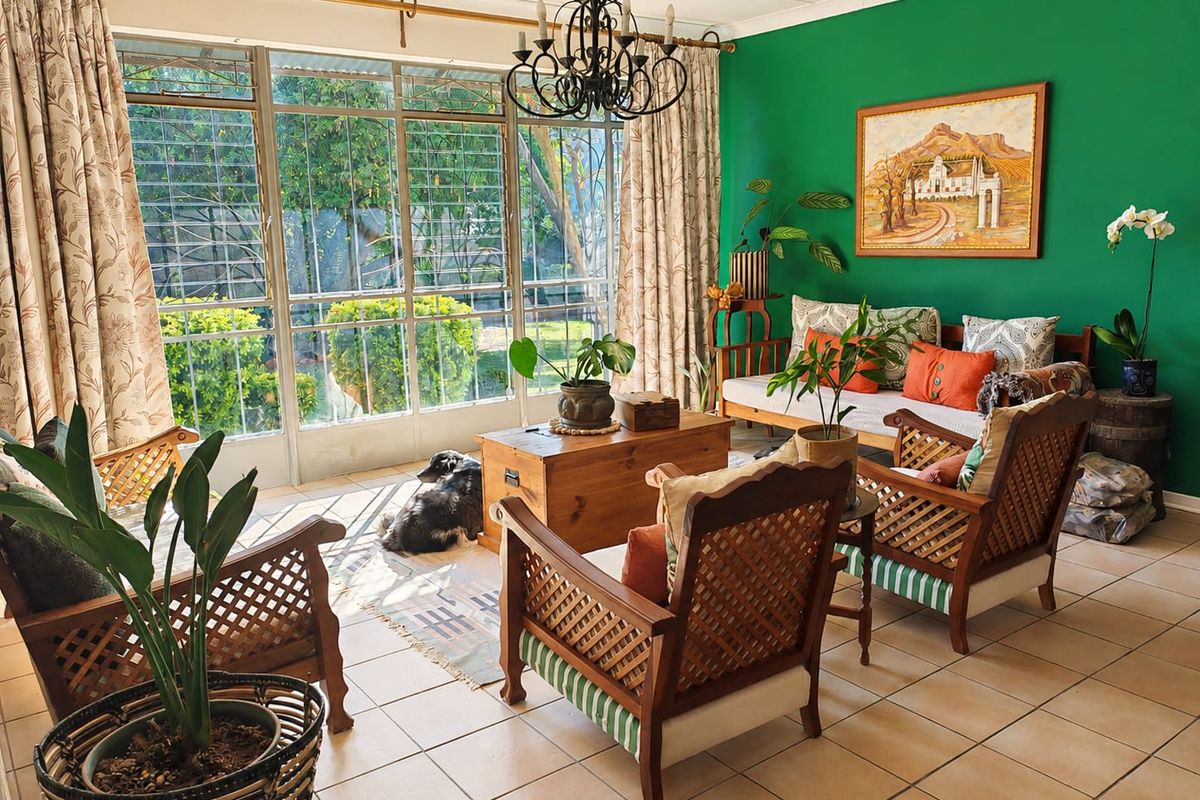 A bright living room featuring tiled flooring, a decorative wrought iron chandelier, and large metal-framed windows with security bars overlooking the garden.