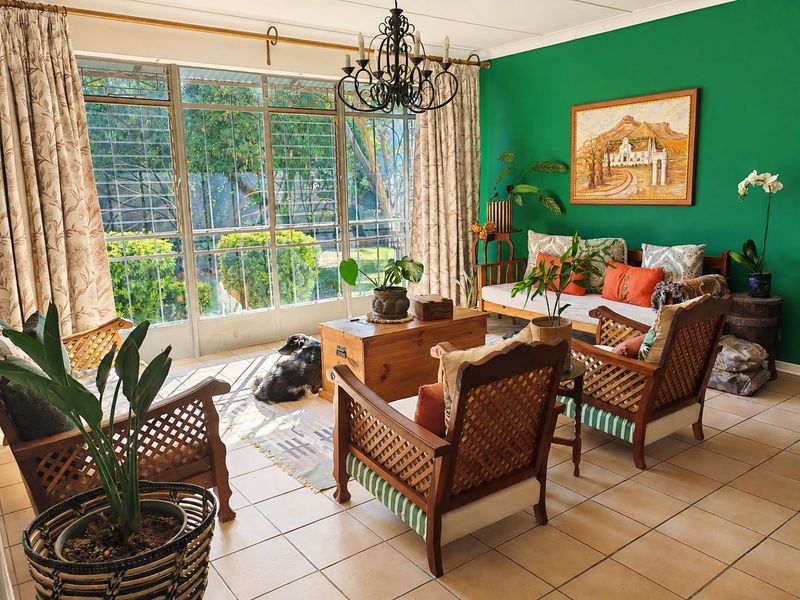 A bright living room featuring tiled flooring, a decorative wrought iron chandelier, and large metal-framed windows with security bars overlooking the garden.