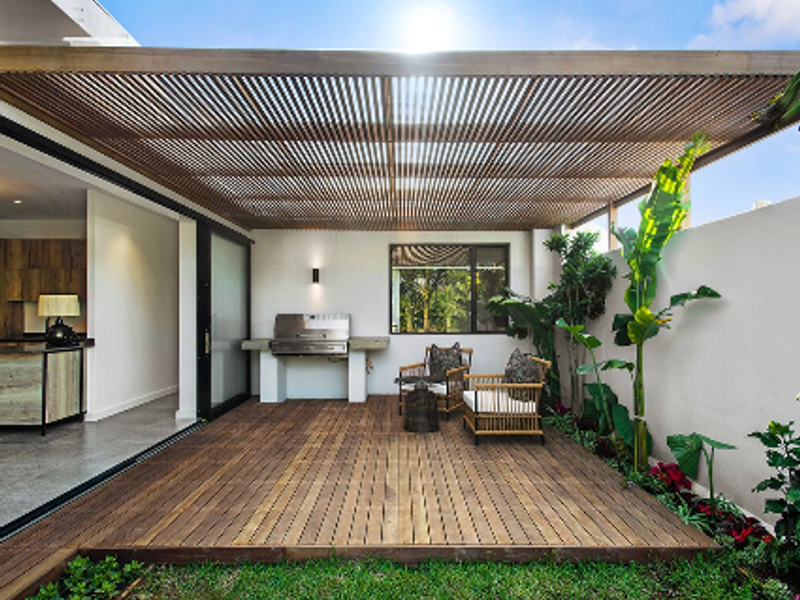 A modern covered patio featuring timber decking, a slatted pergola, and a built-in barbecue, seamlessly connecting to the interior living space via sliding doors.