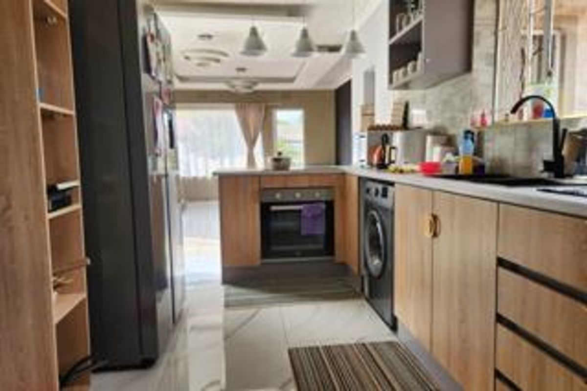 A functional kitchen featuring light wood-finish cabinetry, built-in oven, integrated laundry appliance, and tiled flooring, leading into an open-plan living area.